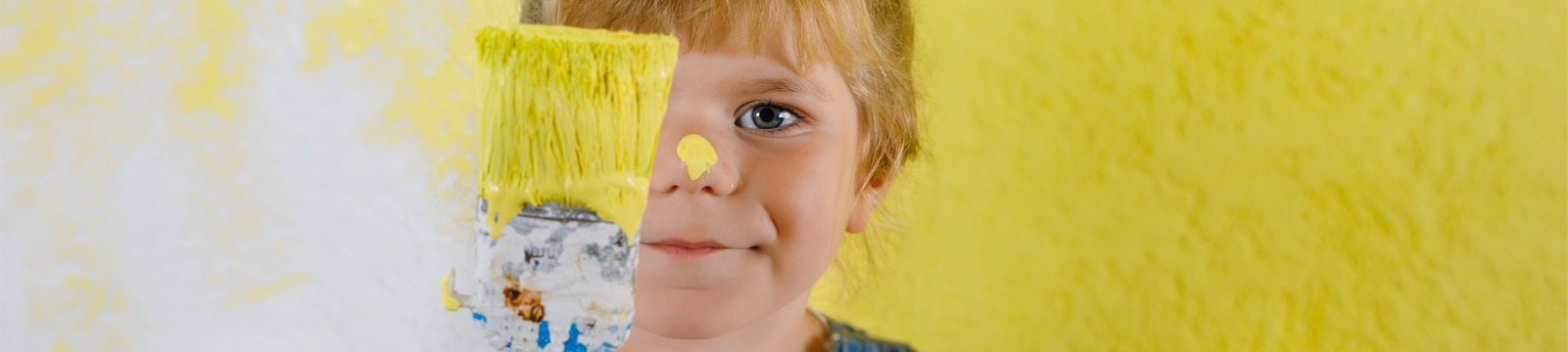 image of cute young blond girl "helping" paint a room a bright, cheerful yellow. She's holding a paintbrush dripping down her arm. Has a big splotch of yellow on nose.